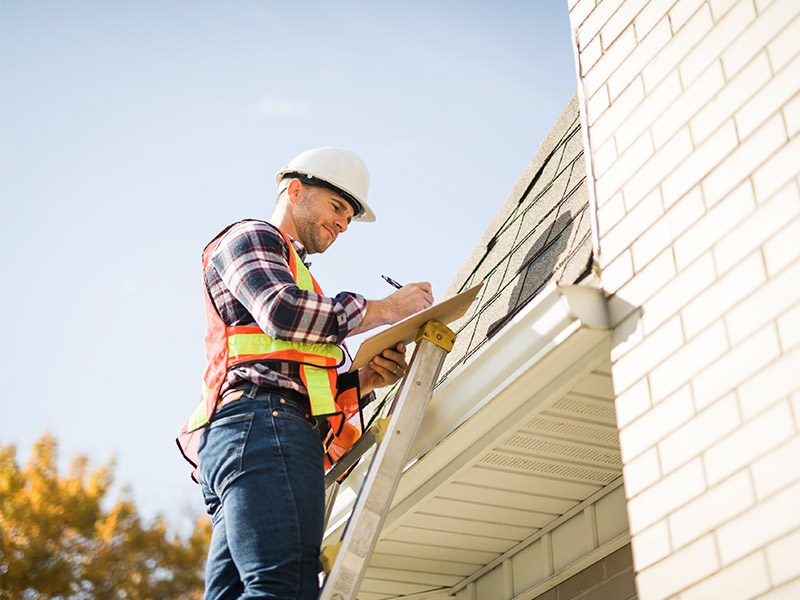 Roofer standing on a ladder inspection roof and writing down findings.